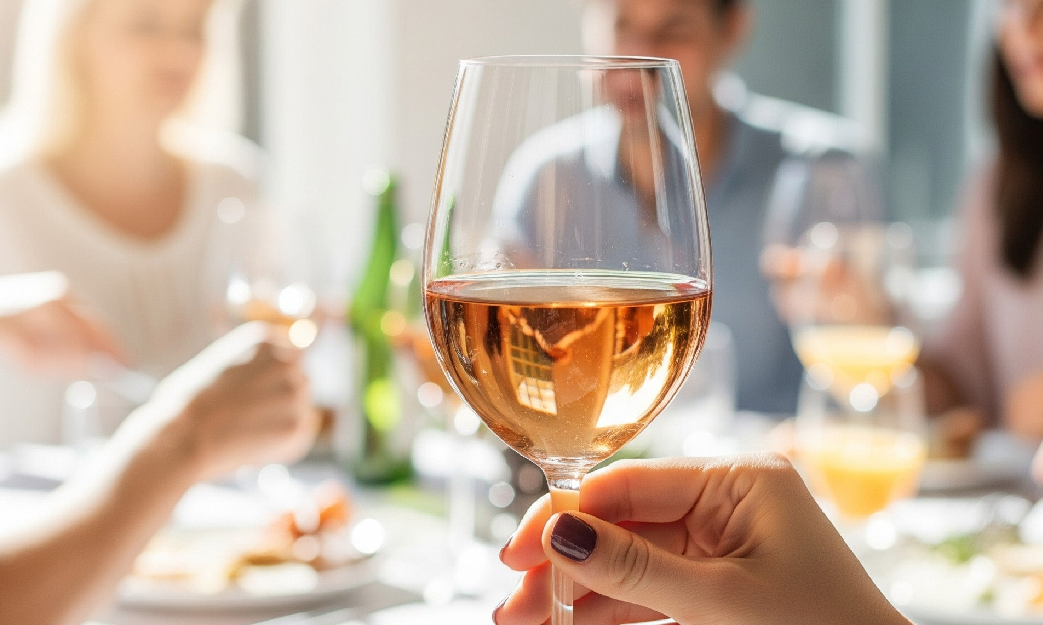 A person&#39;s hand holds a glass of rosé wine, with a blurred background showing people enjoying brunch in a light-filled room.