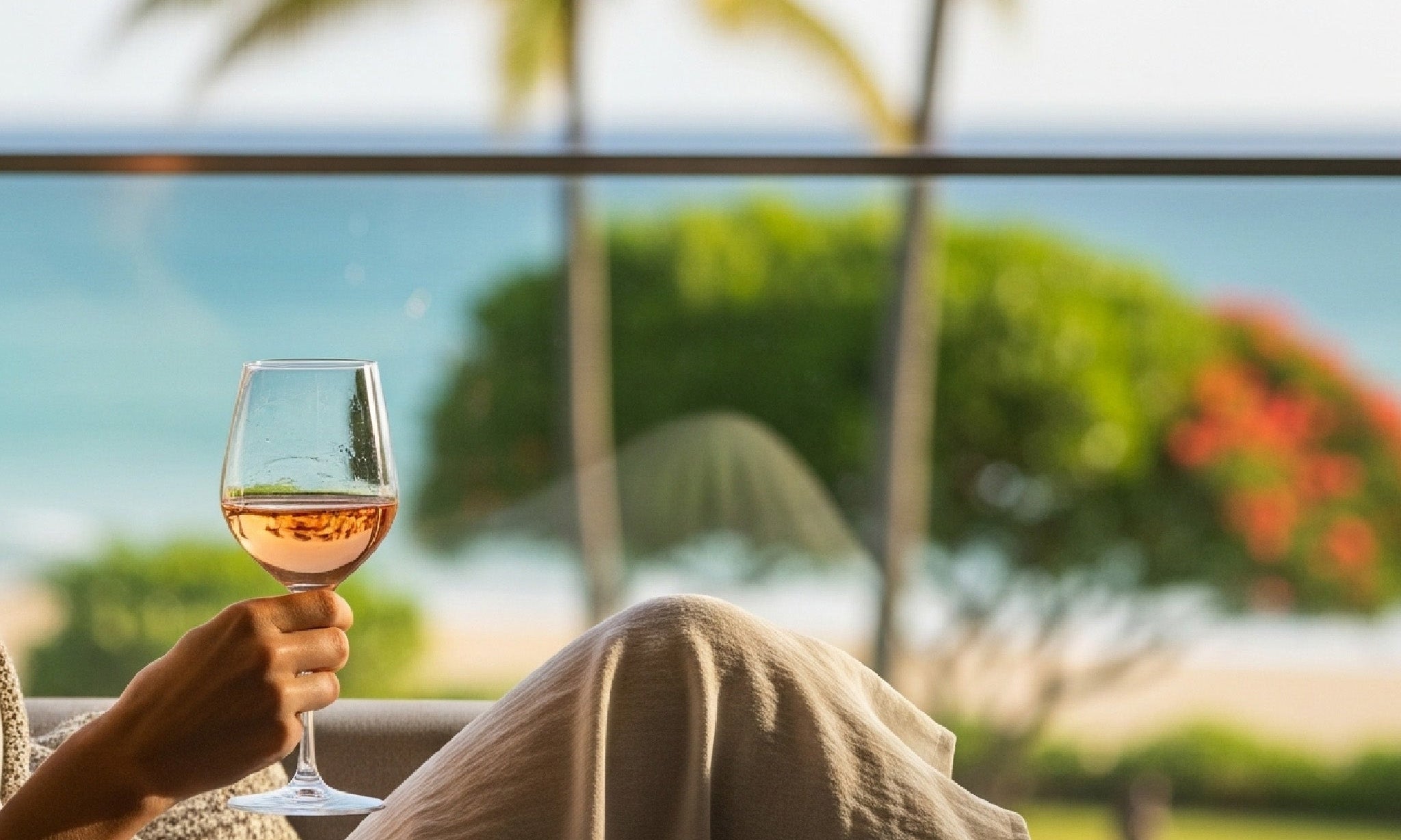 A woman wearing a light-colored dress and a cozy blanket relaxes on a sofa, holding a glass of wine, with a tropical beach and palm trees visible in the background.