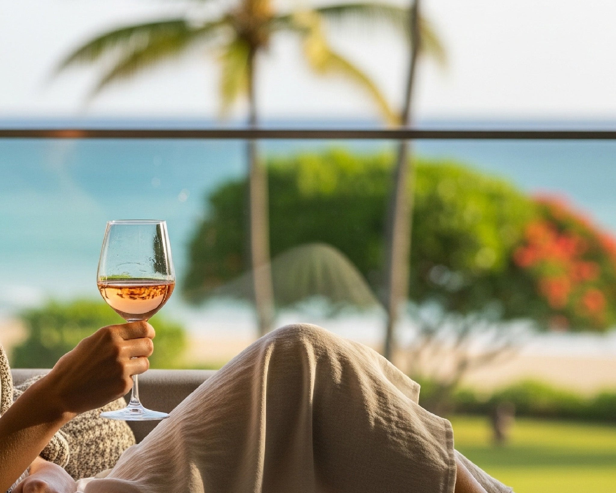 A woman wearing a light-colored dress and a cozy blanket relaxes on a sofa, holding a glass of wine, with a tropical beach and palm trees visible in the background.