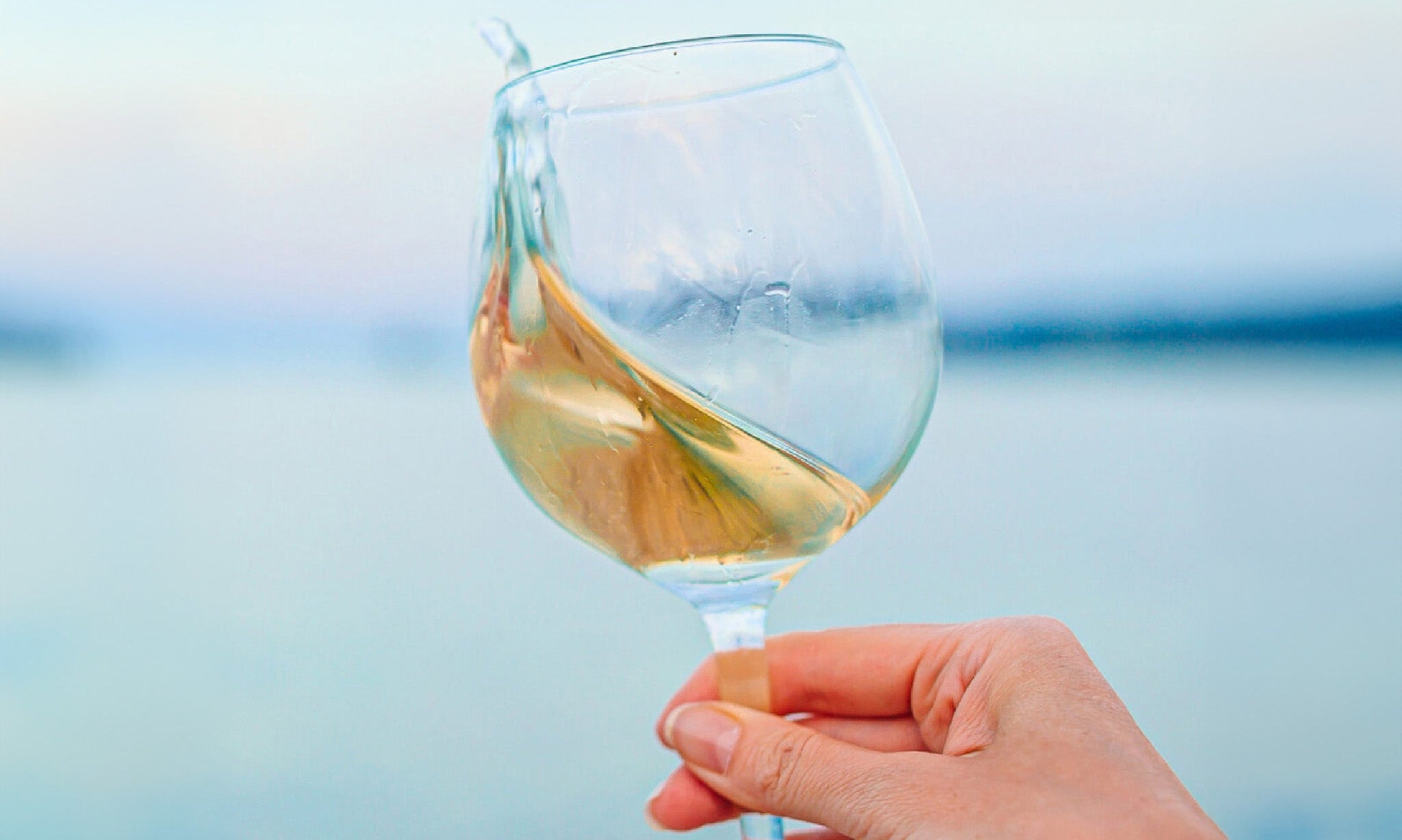 A hand holding a glass of cold white wine with the sea in the background.