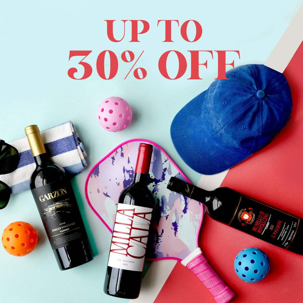 A promotional flat-lay featuring two bottles of red wine, a blue baseball cap, a pink-and-white paddle, a striped towel, and colorful balls on a split light blue and red background, with large text reading 'UP TO 30% OFF'.
