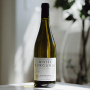 A bottle of Berry Brook & Rudo White Burgundy wine stands on a light surface with a softly blurred indoor background featuring warm sunlight and greenery.