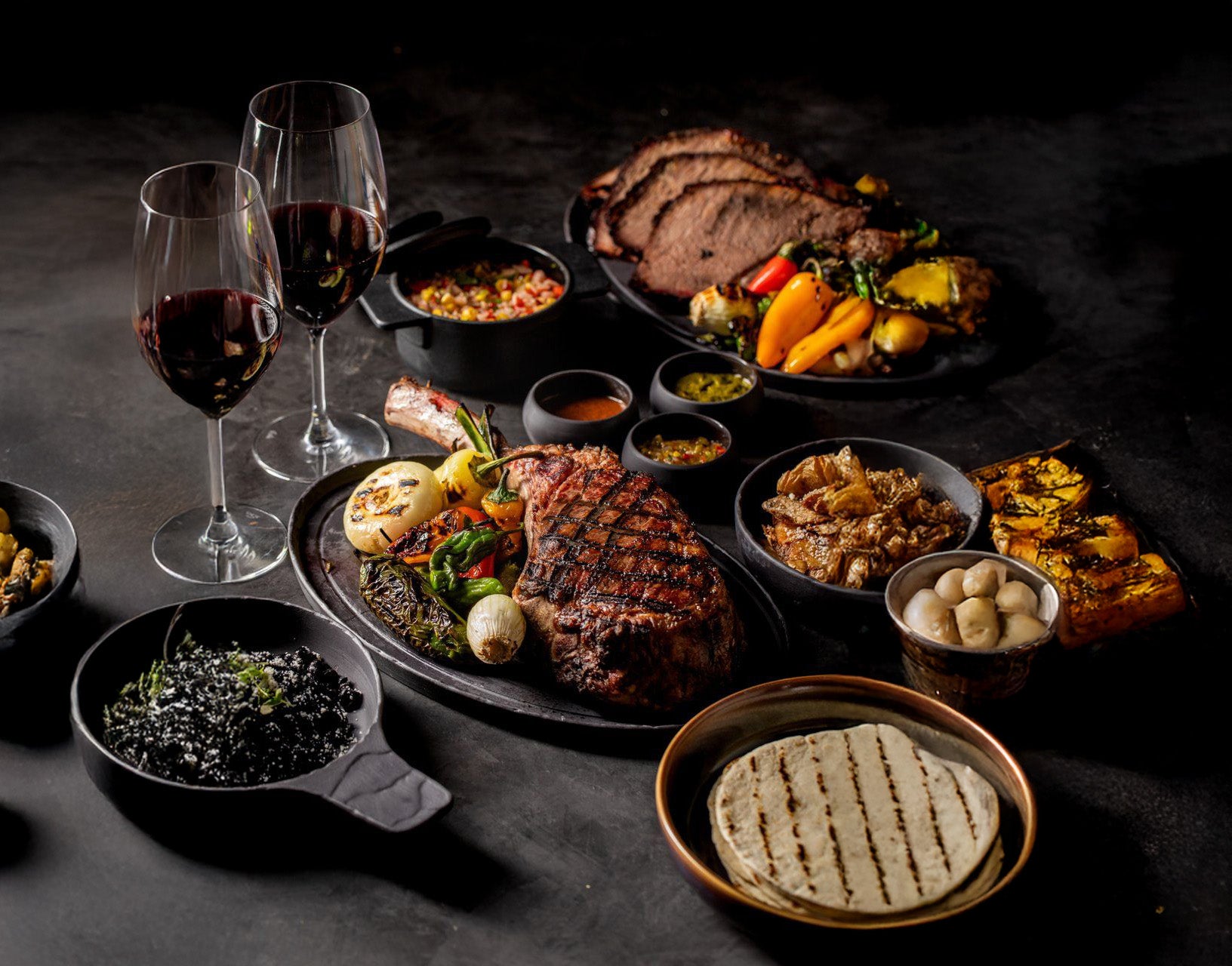 Assorted meats, vegetables, and wine glasses on a dark surface