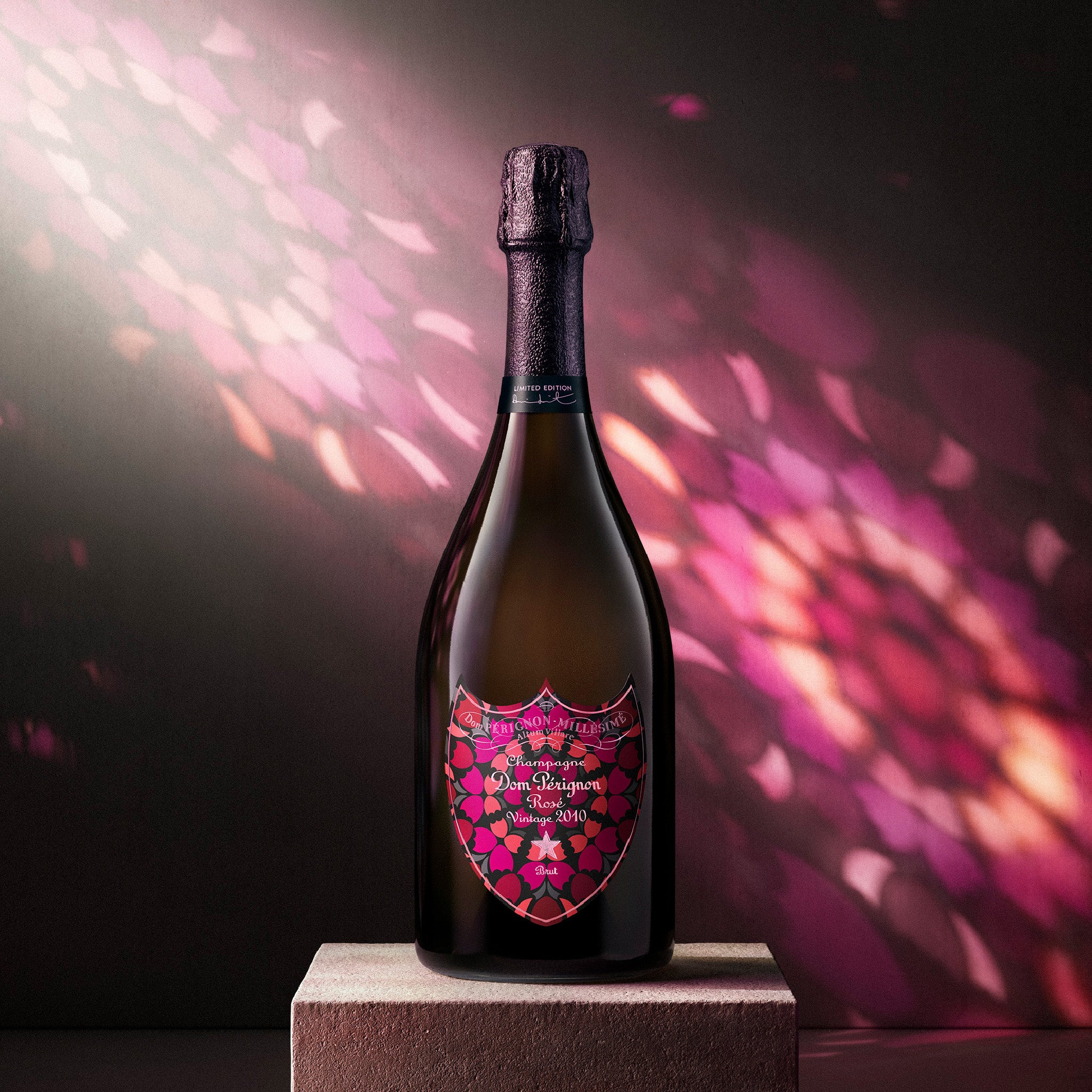 A bottle of Brut Champagne with a custom pink and orange mandala-patterned label, displayed on a stone pedestal against a dark background with pink bokeh light effects.