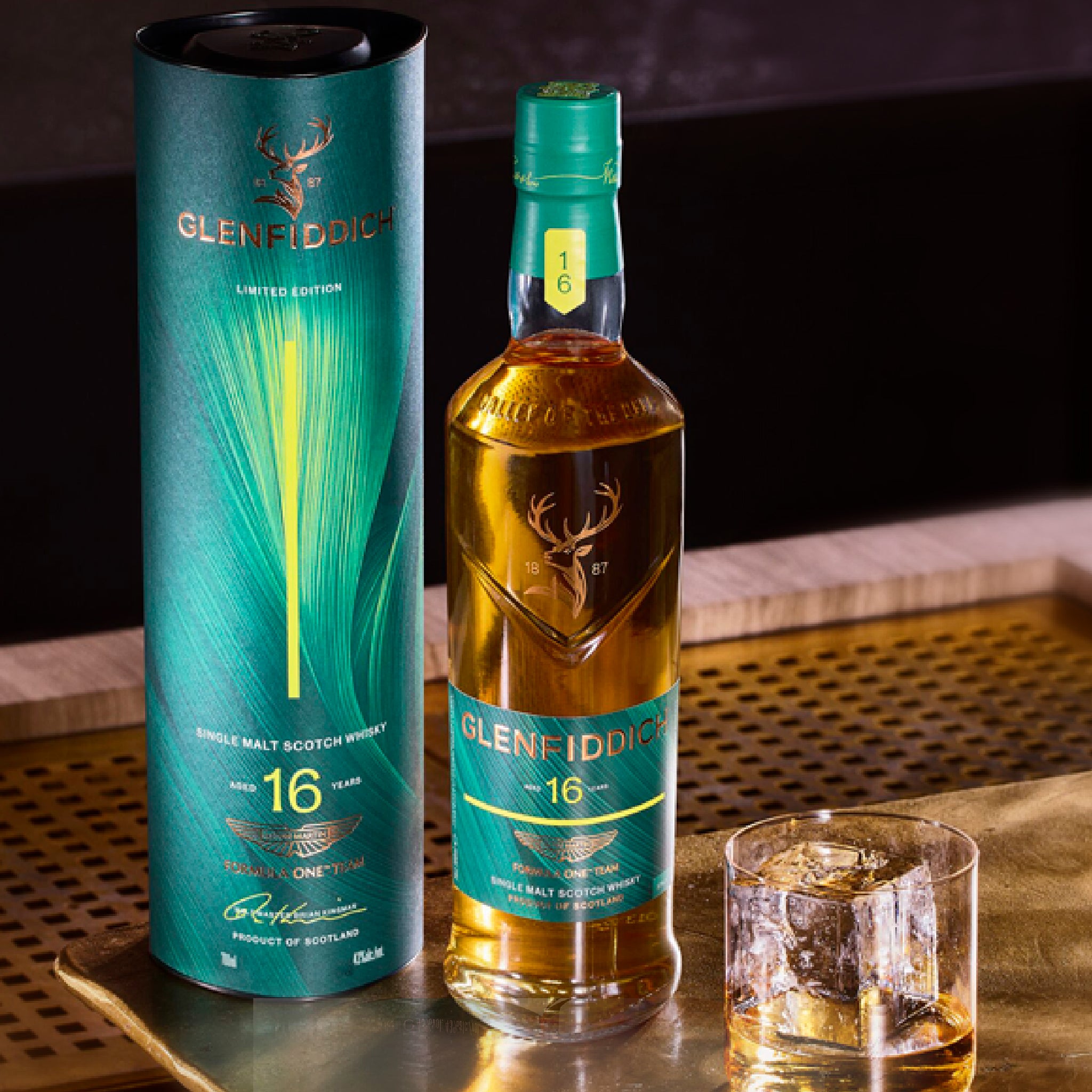 A bottle of Glenfiddoid 16 Year Single Malt Scotch Whisky stands next to its teal cylindrical packaging tube on a wooden tray, with a filled rocks glass in the foreground.