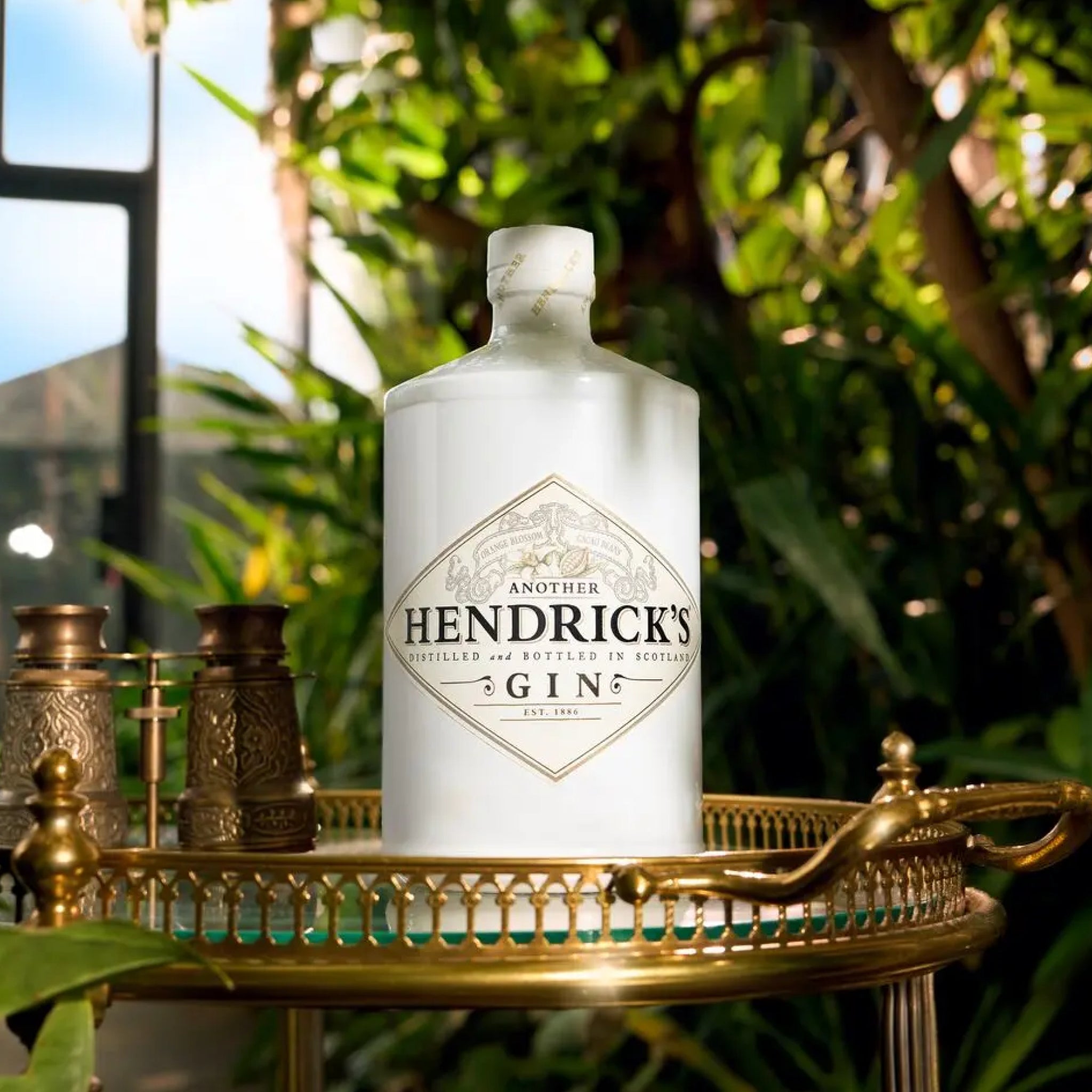 A white bottle of Hendrick's Gin stands on a gold-toned tray in a lush green outdoor setting with foliage and a window in the background.
