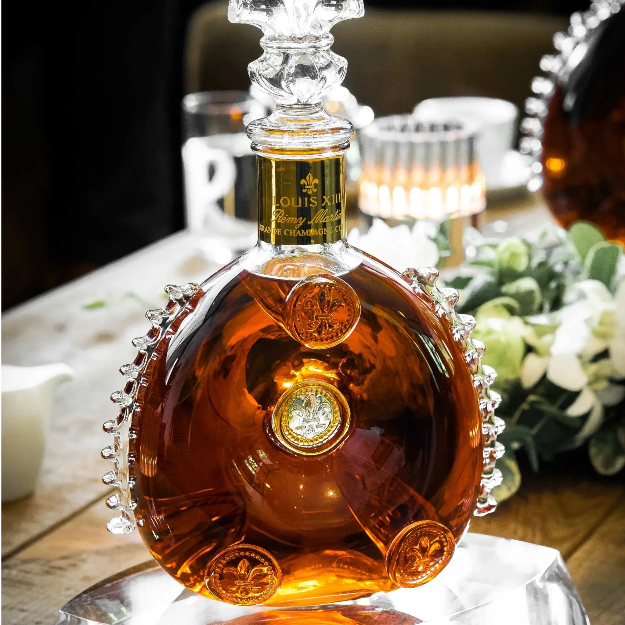 A Louis XIII cognac bottle filled with amber liquid, displayed on a table with glasses, flowers, and a softly blurred background.