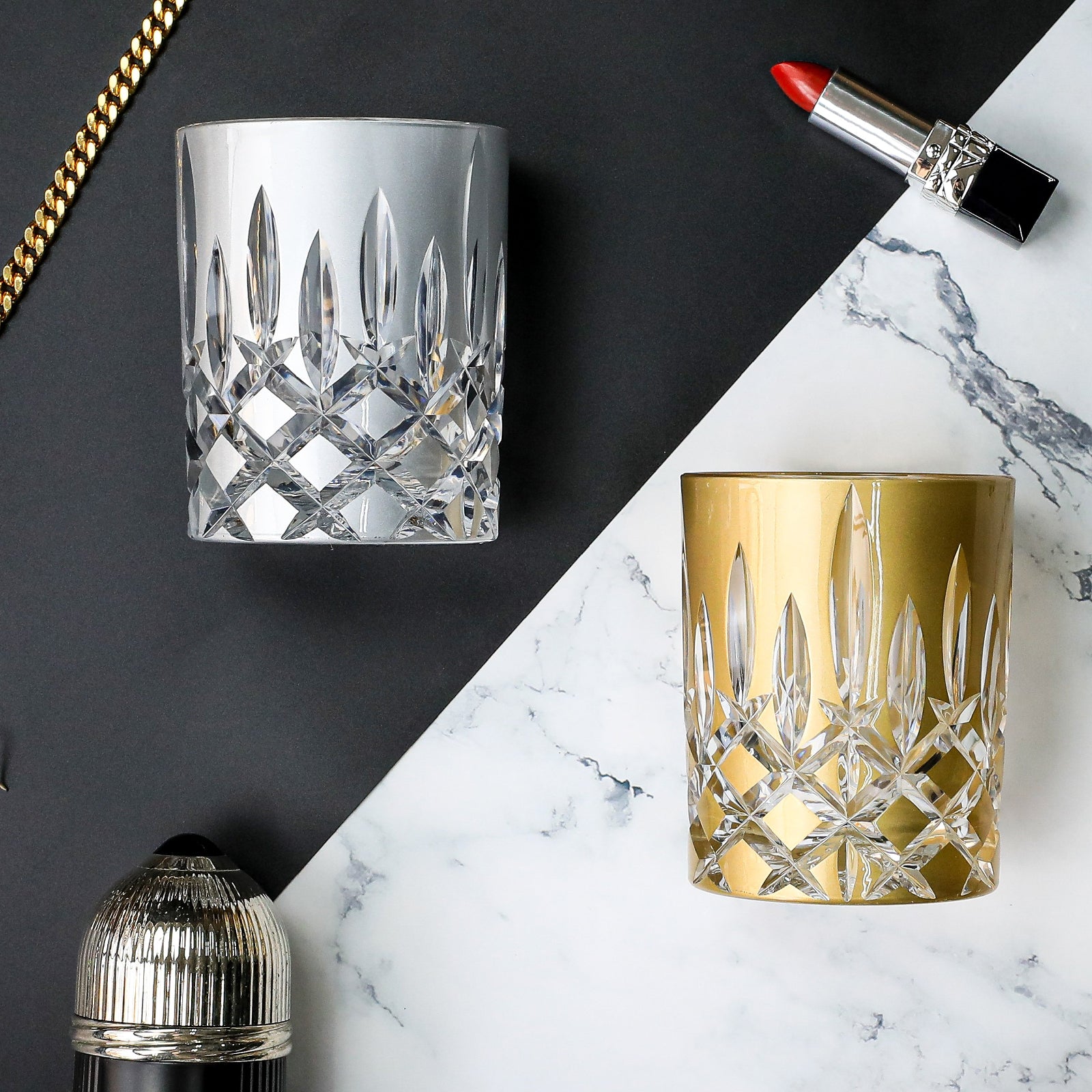 Two crystal glasses, one clear and one gold, on a black and white marble surface with a lipstick and lip gloss.
