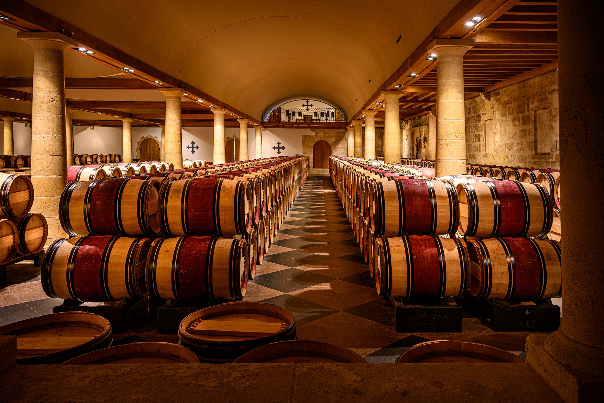 Wine barrels arranged in rows inside a large wine cellar with stone walls and high ceilings.